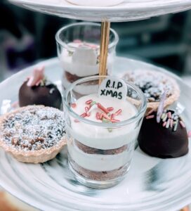 Scary Merry Christmas includes a photo of a plate with small desserts including a chocolate truffle with festive sprinkles, a small tart with powdered sugar and a layered trifle in a small glass with cake, frosting and colorful sprinkles with a decoration saying R.I.P Xmas