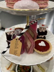 A three-tiered serving tray filled with an array of delicious treats. The bottom tier holds finger sandwiches and savory bites. The middle tier features a "Drink Me" potion bottle, a pink cake slice, and a heart-shaped tart. The top tier displays delicate pink macarons and a whimsical cake decorated with a playing card.